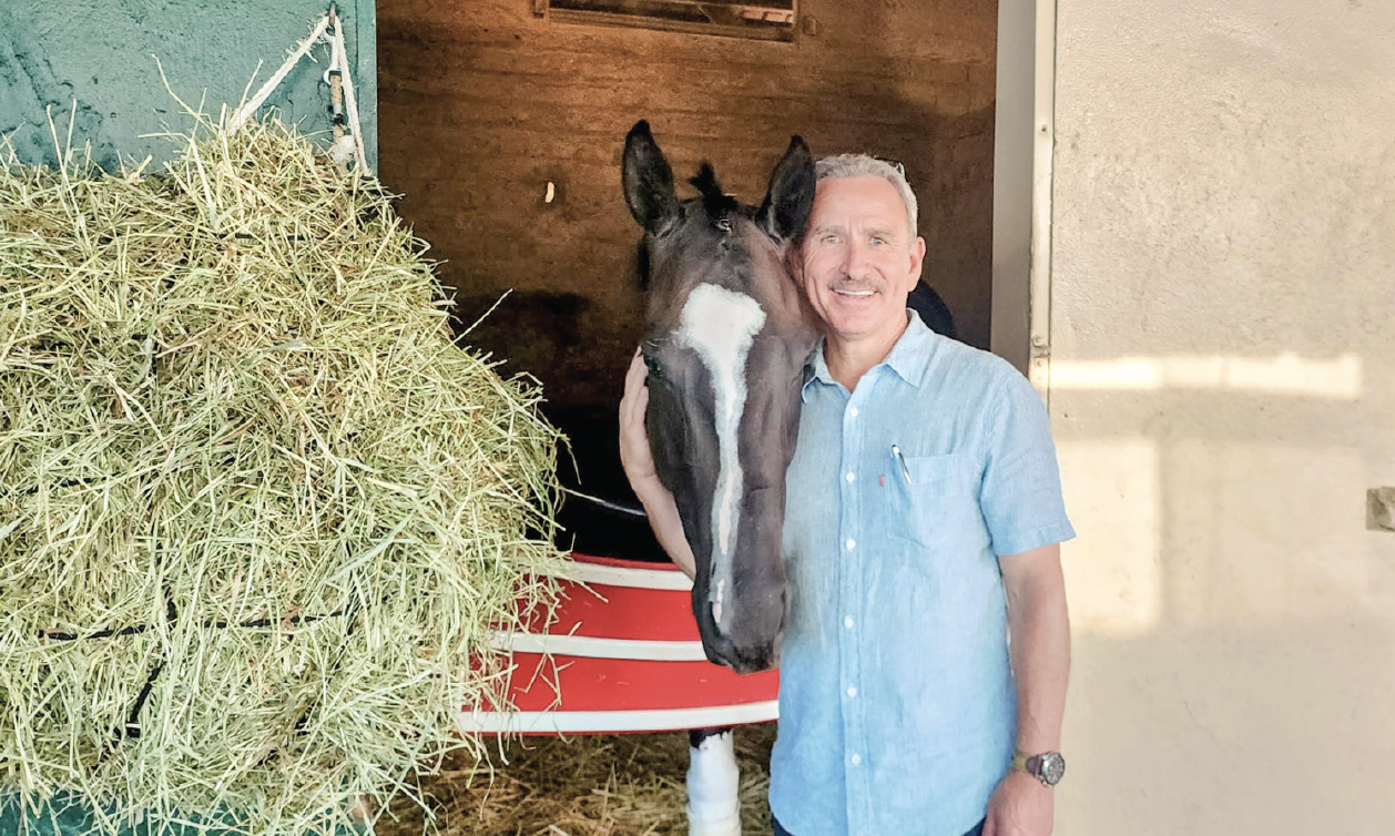 A smiling man in a light blue shirt stands beside a dark brown horse with a white stripe on its face. They are in a stable doorway with hay stacked to the left.